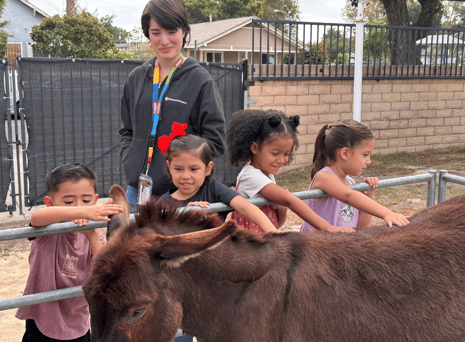 Young students smiling and petting a donkey over a fence while a staff member wearing a lanyard supervises the activity outdoors.