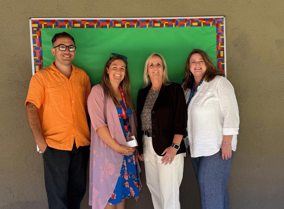 Four Santa Ana Classical Academy staff members posing together in front of a green bulletin board outdoors.
