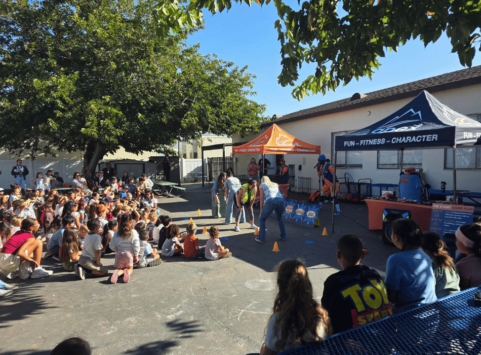 A large group of students sitting outdoors on asphalt watching staff members participate in an Apex Leadership event set up with event tents.