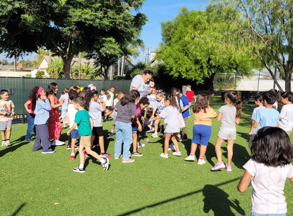 Elementary students gathered outside on a green turf field participating in organized group games with a staff member.