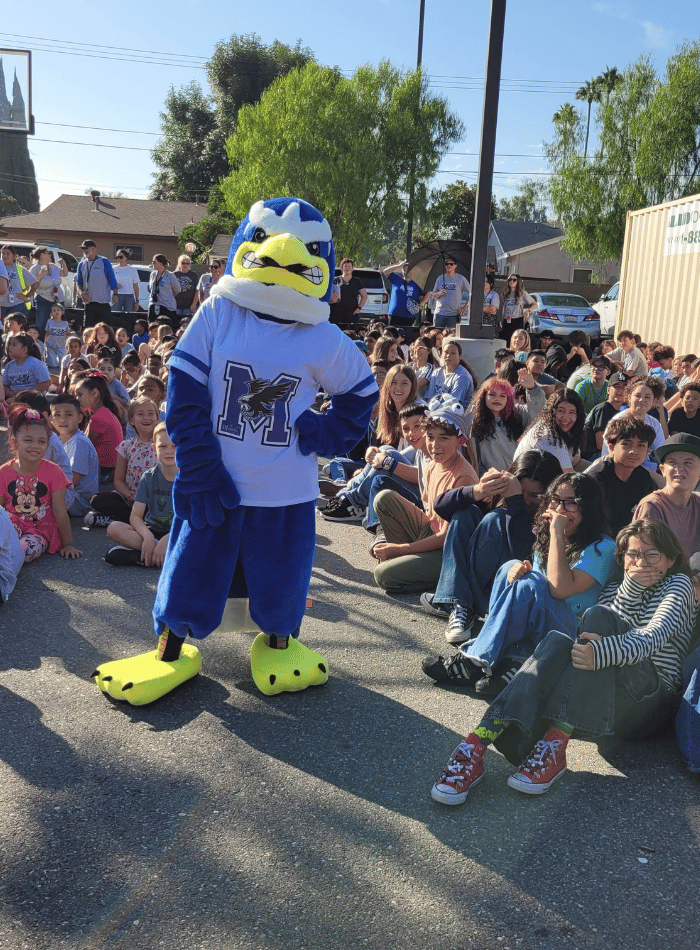 A blue and white bird mascot standing in front of a large group of students seated outdoors on the asphalt during a school assembly.