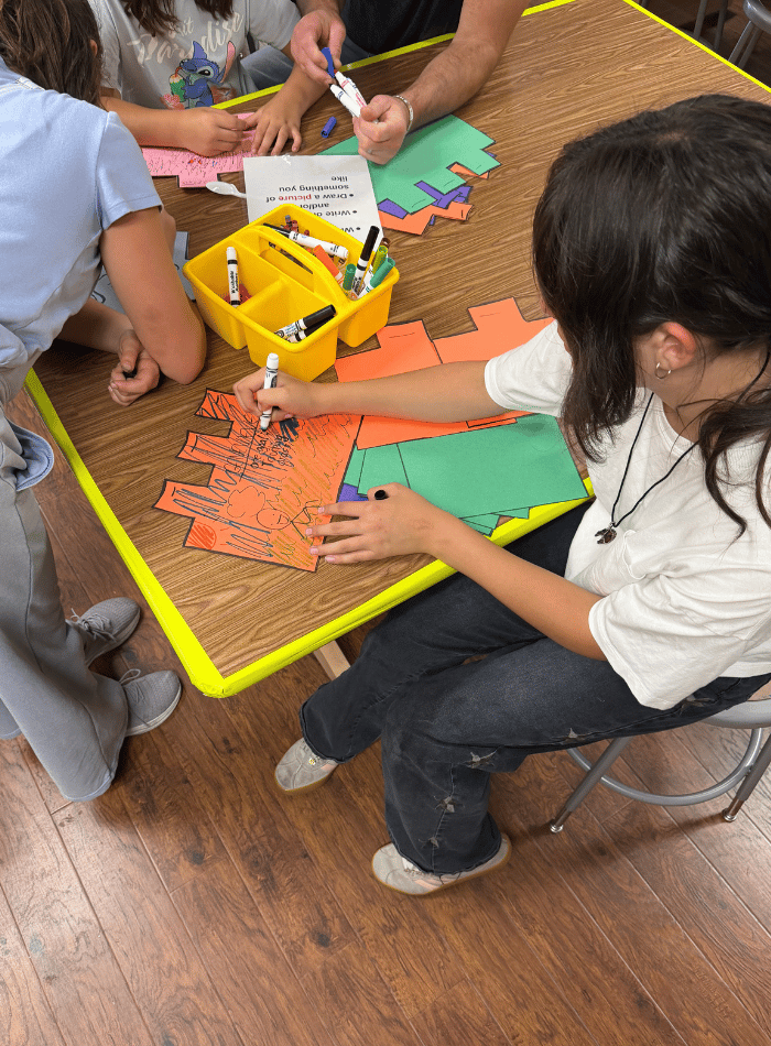 Students sitting around a classroom table using markers to color and write on block-shaped construction paper cutouts during Back to School Night at Discovery Charter School.