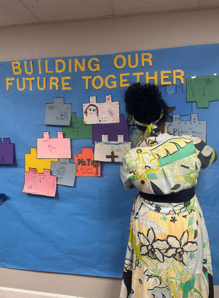 A teacher hanging student-decorated paper building blocks onto a blue bulletin board titled "Building Our Future Together" during Back to School Night at Discovery Charter School.
