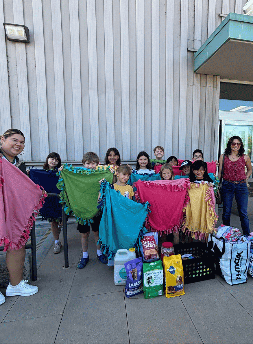 Students and staff standing outside an Animal Services building holding up colorful handmade blankets next to donated pet food and supplies