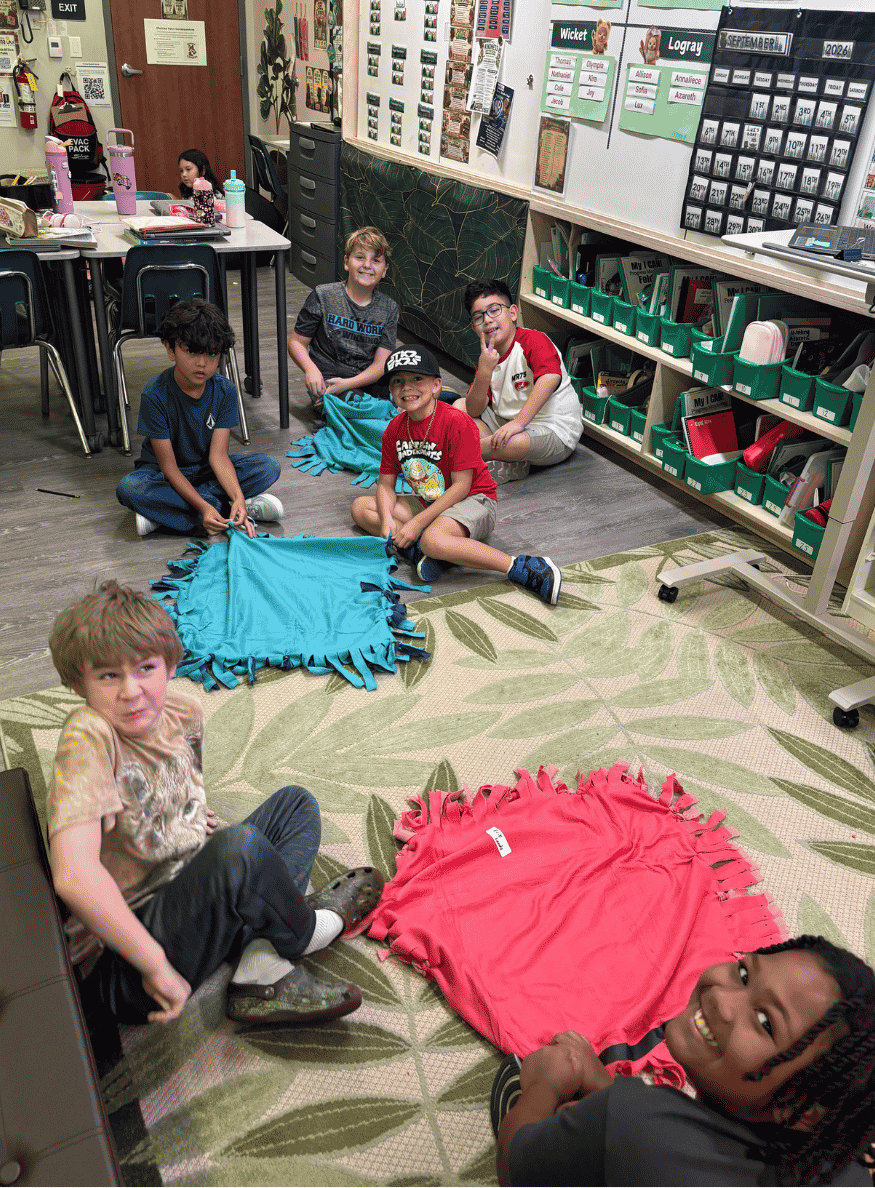 Elementary students sitting on the floor in a classroom assembling colorful tie-fleece blankets for a service project.