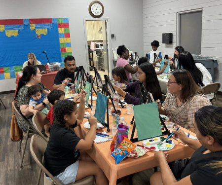 Students and family members sitting around tables with easel canvases and paint sets during Del Rio Paint Night.
