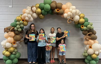 Students and parents posing under a decorative balloon arch while holding up their finished sunflower canvas paintings at Del Rio Paint Night.