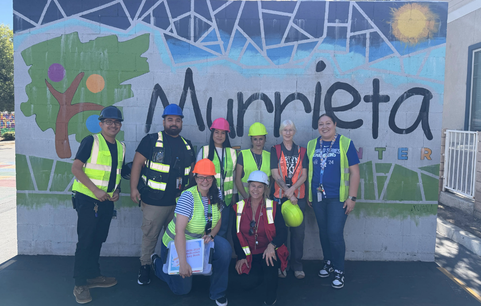 Murrieta Student Center staff members wearing hard hats and high-visibility safety vests posing together in front of the school mural.