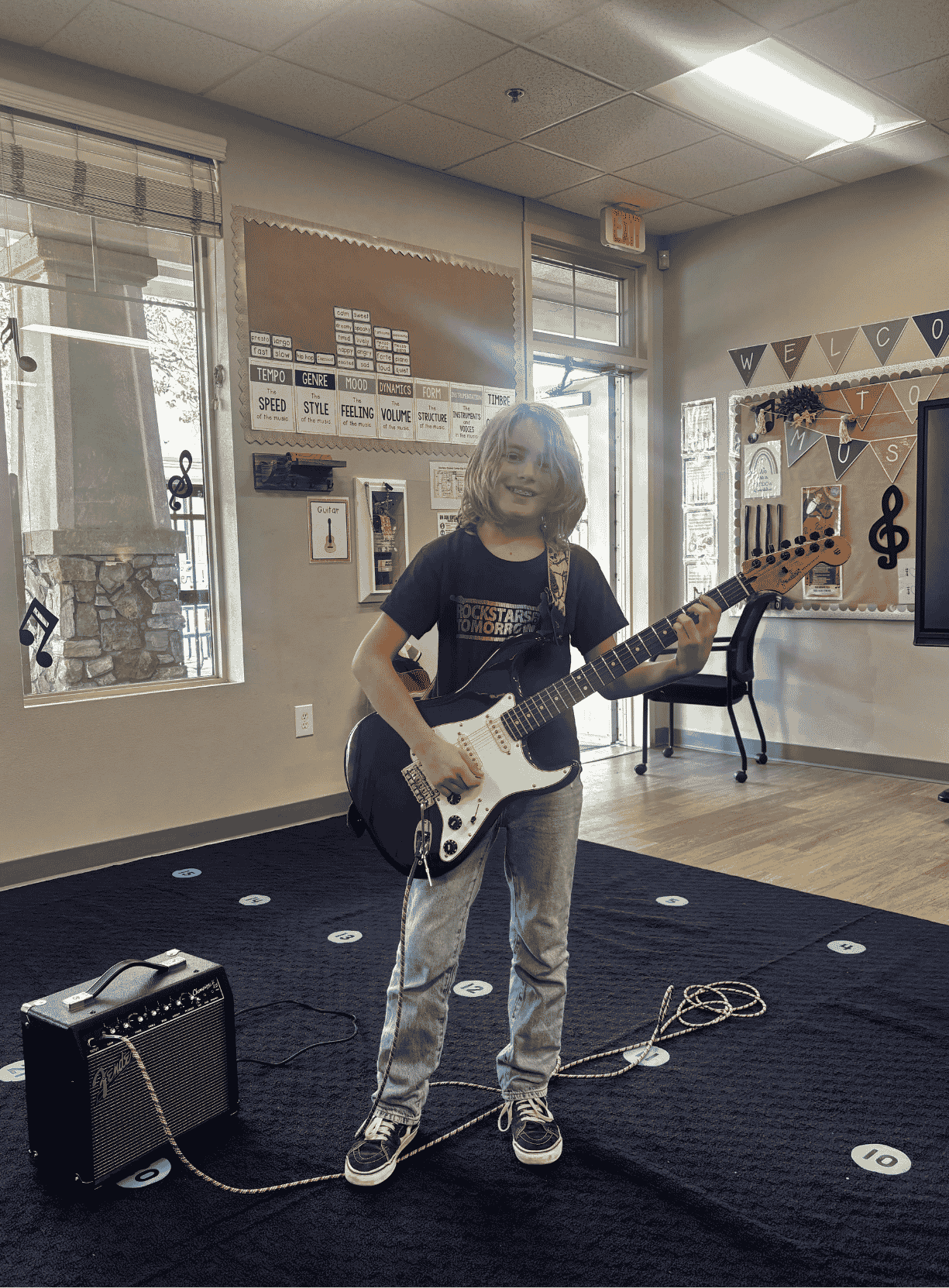 A student standing in a music room holding an electric guitar plugged into an amplifier.