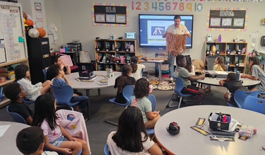 A teacher standing in front of a classroom displaying a fringed artifact to students seated at round tables during a lesson at Otay Ranch Academy.