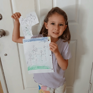 A young student smiling while holding up her hand-drawn family portrait and star drawings on lined paper.