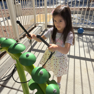 A young student holding mallets to play a green outdoor bell chime instrument on the Temecula Student Center playground