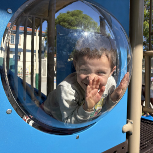 A young student smiling and pressing his hands against a clear bubble window on a blue playground structure.