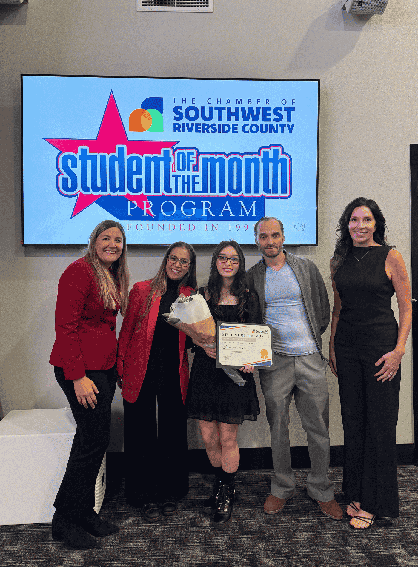 A Journey Homeschool student holding a bouquet and certificate while posing with family and staff in front of a Student of the Month screen.
